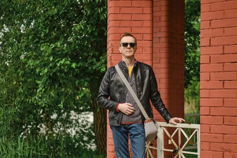 A man stands with a relaxed posture beside vibrant red brick pillars, dressed in a casual outfit