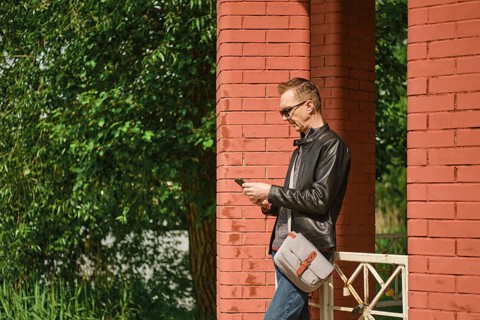 A man wearing sunglasses and a leather jacket stands by red brick columns, attentively using his
