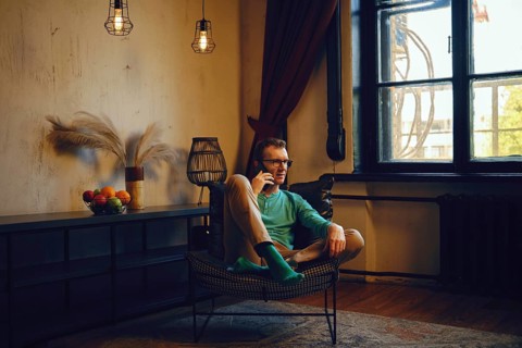 A man dressed in a green long-sleeve shirt and light pants relaxes in a chair, engaging in a phone