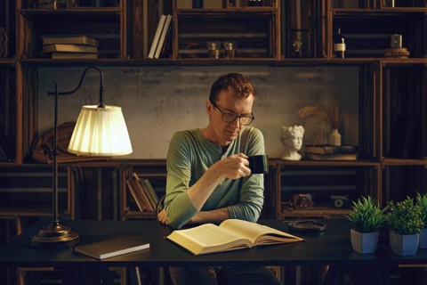 A middle-aged man sits in the dimly lit room in the evening and reads an exciting book. He drinks