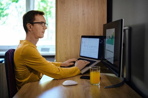 A middle-aged man focused on his work sits at a wooden desk with two computer screens. He enters