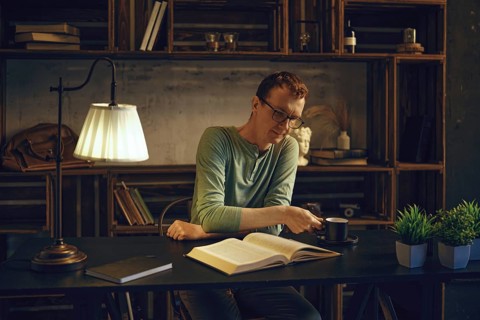 A man enjoys a quiet evening in a rustic home office, reading a book while sipping herbal tea. The