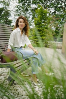A woman with curly hair relaxes on a wooden bench in a serene park setting, wearing a white shirt