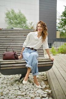 A woman sits comfortably on a modern bench in a lush park, wearing a light button-up shirt and