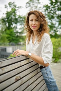 A woman with curly hair stands confidently by a wooden railing in a green park. She wears a light