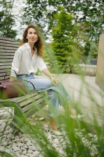 A woman sits calmly on a wooden garden bench, dressed in a light shirt and jeans. Tall grass sways