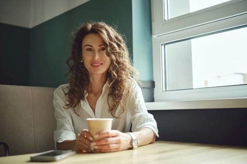 A woman with curly hair sits at a wooden table in a cafe, holding a cup of coffee. She smiles while