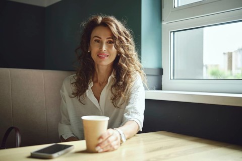 A woman with curly hair sits at a table near a window, holding a cup of coffee. She wears a white