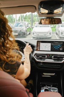 A woman with curly hair is focused on driving a car, checking the navigation screen. She is inside