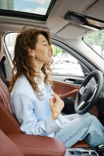 A woman with curly hair is seated in the driver's seat of a modern car, smiling and looking forward