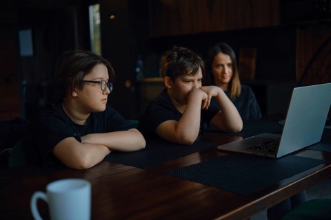 Two children and their mom watching movie on a laptop. They sit at a wooden table in a comfortable