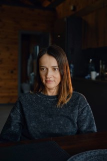 A woman with medium-length brown hair sits at a dark wooden table, reflecting thoughtfully. The