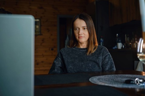 A woman is focused on her laptop while sitting at a wooden table in a warm dark living room. The