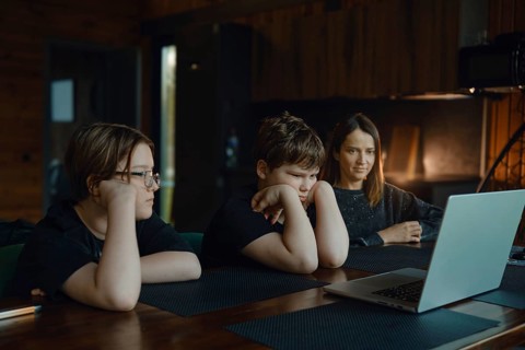 Two children and their mother sit at a wooden table, watching a laptop screen. The room is warm and