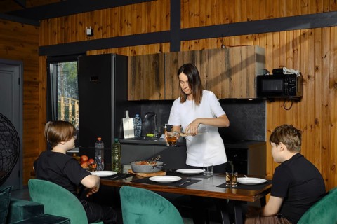 Two children sit at a dining table while their mother pours drink in a cozy cabin kitchen. A pan