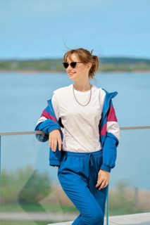 A woman stands confidently by the water, dressed in comfortable sportswear and sunglasses. She