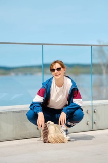 A young person squats on a modern deck by a scenic body of water, wearing sunglasses and a sporty