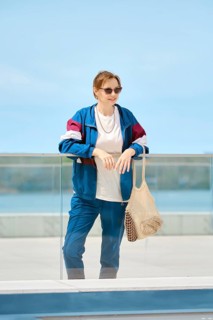 A woman stands by a glass railing at a waterfront location, dressed in a colorful jacket and