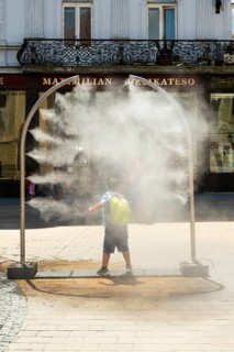 A mobile spray arch sprays a fine mist of water in a sunny square. A child uses the refreshment in
