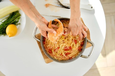 A person is adding salmon to a pot of pasta with tomatoes and herbs on a sunny kitchen countertop.