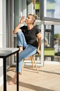 A middle-aged woman sits comfortably on a wooden chair in a sunny balcony, sipping from a glass.