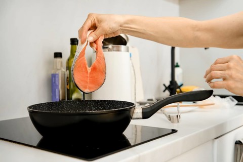 A hand is lifting a fresh salmon fillet above a black frying pan on a stovetop. The kitchen appears