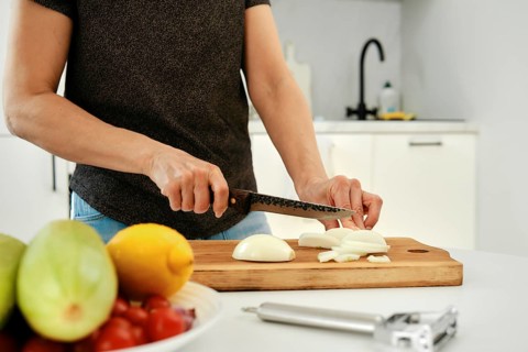 A person is slicing fresh onion on a wooden cutting board in a sleek kitchen. Colorful fruits and