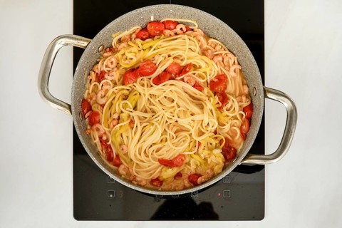 A pot of spaghetti is simmering on a stove, mixed with shrimp and cherry tomatoes