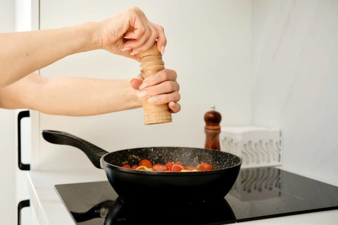A hand is grinding pepper over a skillet filled with fresh tomatoes. The setting is a modern