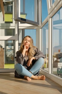 A middle-aged woman sits comfortably on a wooden floor of balcony in a bright modern apartment. She