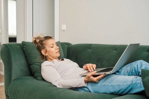 A woman with a relaxed demeanor is laying on a green couch in a bright living room, focused on her