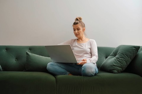 A woman sits cross-legged on a soft green couch, focused on her laptop. Natural light fills the