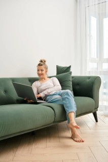 A woman enjoys her time on a green couch, focused on her laptop. Sunlight fills the bright living