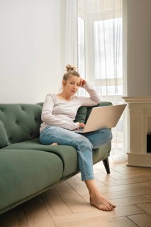 A woman is focused on her laptop while seated on a soft green sofa. She appears thoughtful, with