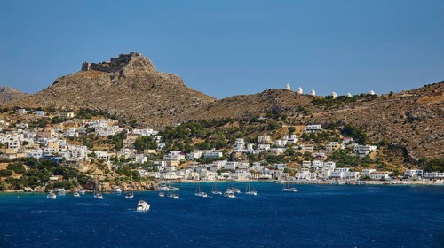 Coastal town on a blue sea with hill and castle in the background, lined with sailing boats,