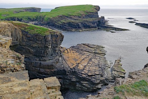 Dramatic rocky coastline with green cliffs and calm sea under a cloudy sky, Orkney Islands,