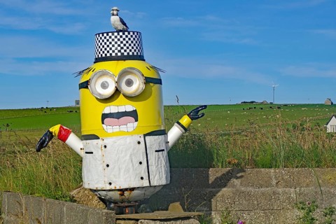 Sculpture of a cartoon figure with a bird on its head, surrounded by a green landscape and blue
