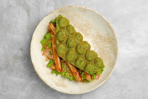 A creatively served sandwich features a green waffle made from spinach, filled with grilled sausage