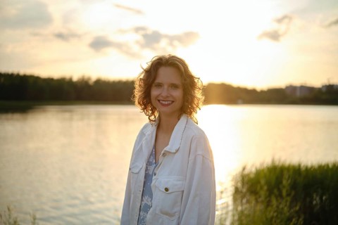 A woman stands near a shimmering lake on a hot evening, her curly hair catching the sunlight. The