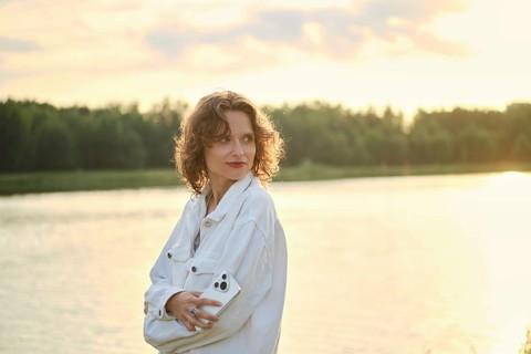 Cute woman with curly hair stands by the lake at sunset. She look back while holding smartphone in