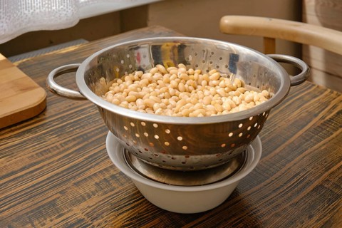 A colander filled with soaked white beans is resting above a bowl to catch excess water. Natural