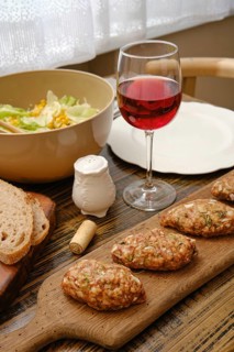 A table is set with a wooden board of homemade patties, a bowl of fresh salad, a glass of red wine,