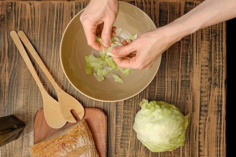 Hands tear crisp lettuce leaves into a bowl, surrounded by wooden utensils and fresh ingredients.