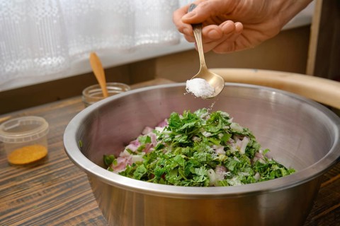 A hand adds salt to a bowl filled with minced meat, chopped onions and fresh herbs. The kitchen is