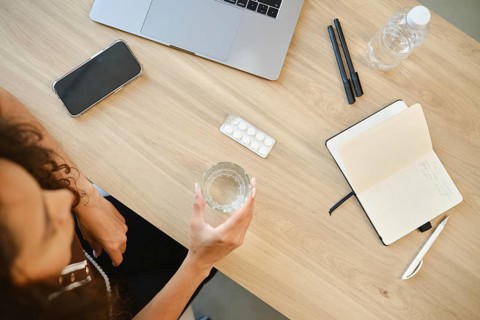 A person sits at a desk with a laptop, holding a glass of water in one hand. A pack of pills and a