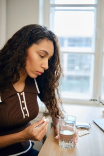 A young woman with curly hair sits at a table concentrating on a pill in her hand and a glass of