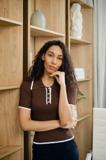 Portrait of a hispanic woman with her hand on her chin, wearing a fitted brown top and black pants.