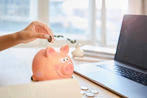 A hand is placing a coin into a pink piggy bank while a laptop sits open on a wooden desk. Sunlight