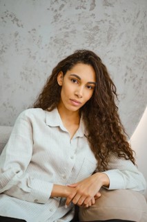 A young woman with long curly hair poses casually in a softly lit modern interior. She rests her