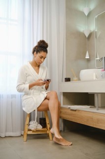 A woman sits on a wooden stool in a contemporary bathroom, wearing a soft white robe. She is